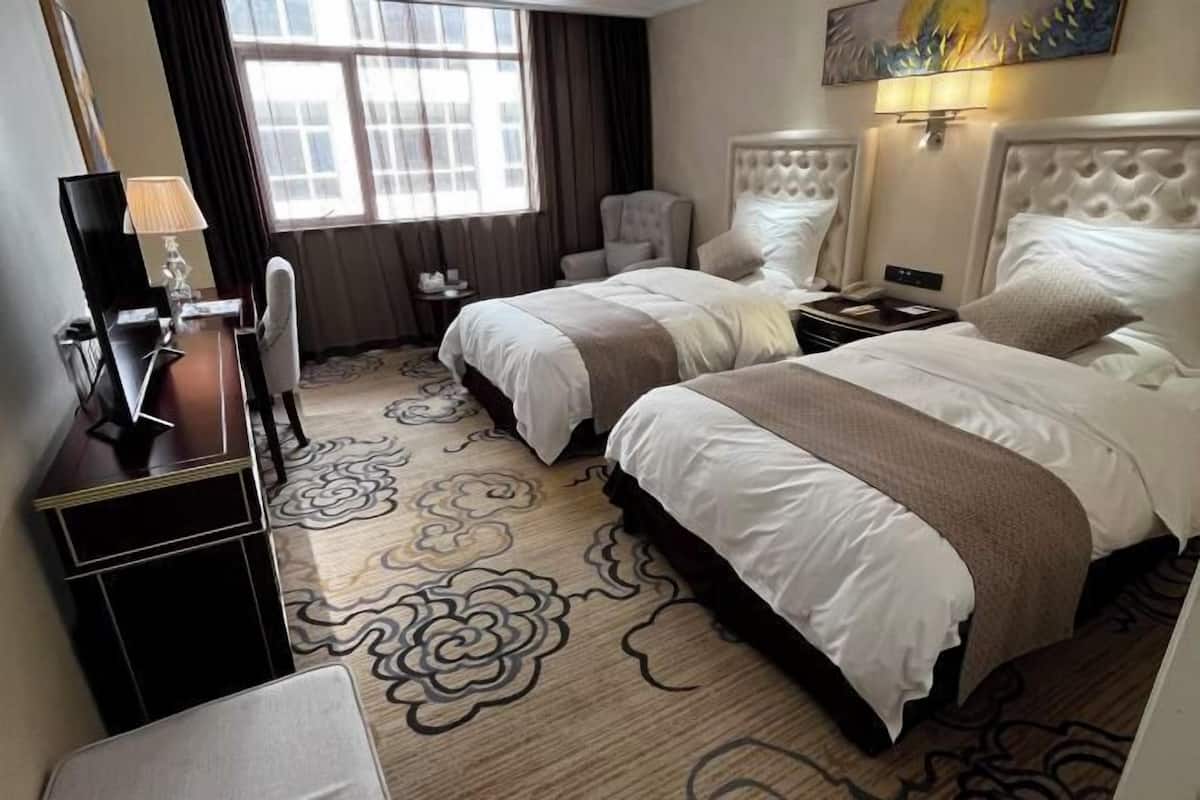 Ein Hotelzimmer mit zwei Betten, einem Schreibtisch mit Lampe, einem Sessel, einem Fernseher und einem großen Fenster mit Vorhängen.
