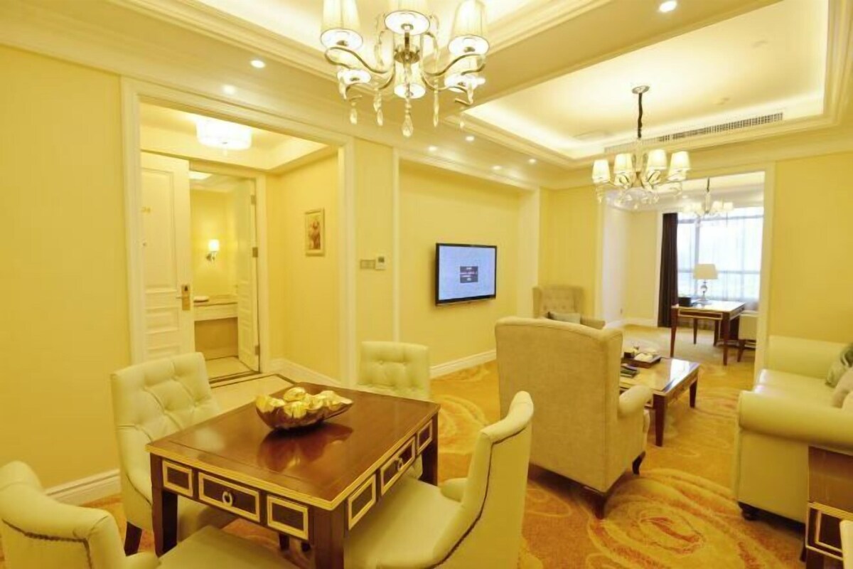 A hotel room with a dining area, a living area with a sofa and armchairs, a TV mounted on the wall, and a chandelier.