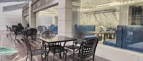 An outdoor seating area with tables and chairs, a covered section with a patterned ceiling, and a view of the interior through a glass wall.