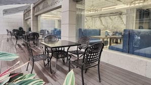 Un área de comedor al aire libre con mesas y sillas, una partición de vidrio y una zona de estar con cojines azules.
