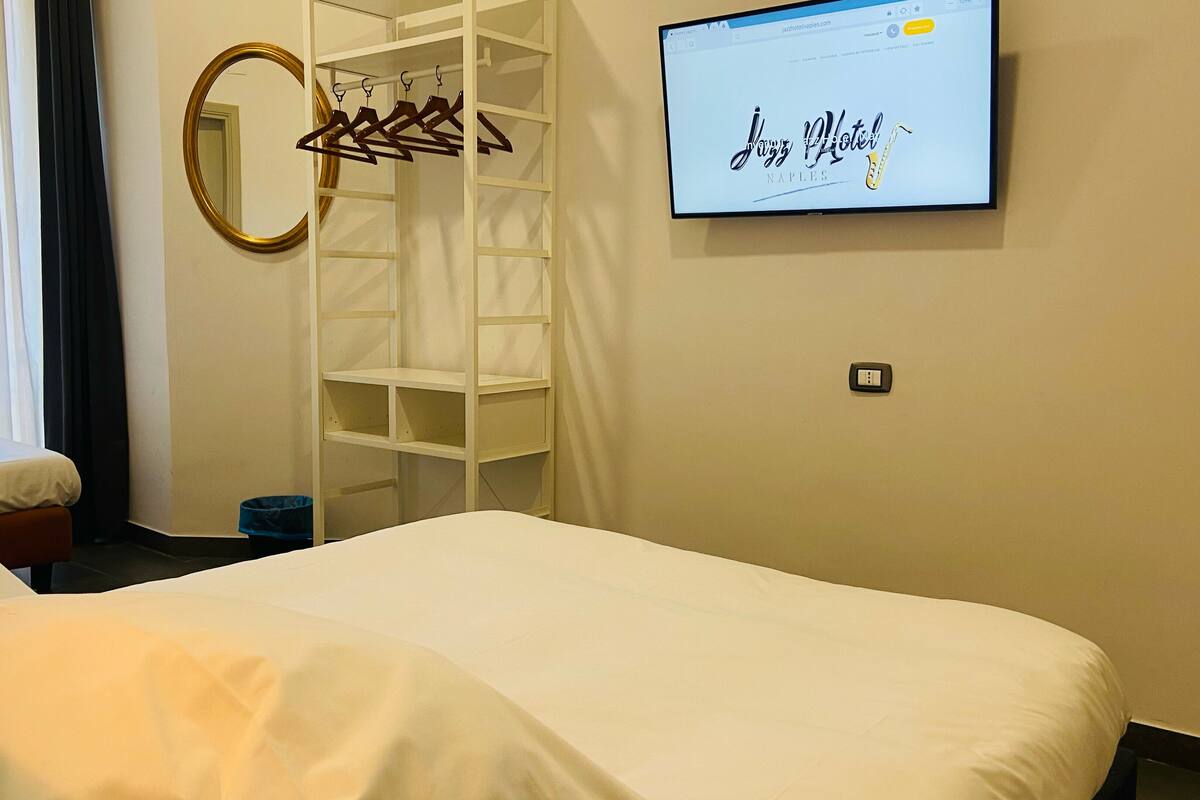 Une chambre d’hôtel équipée d’un lit, d’une armoire, d’un miroir et d’un téléviseur diffusant l’émission « Jazz Hotel Naples ».