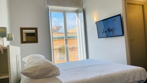 A hotel room with a bed, a window with a view of buildings, a framed picture, and a wall-mounted TV. - Jazz Hotel (Naples)