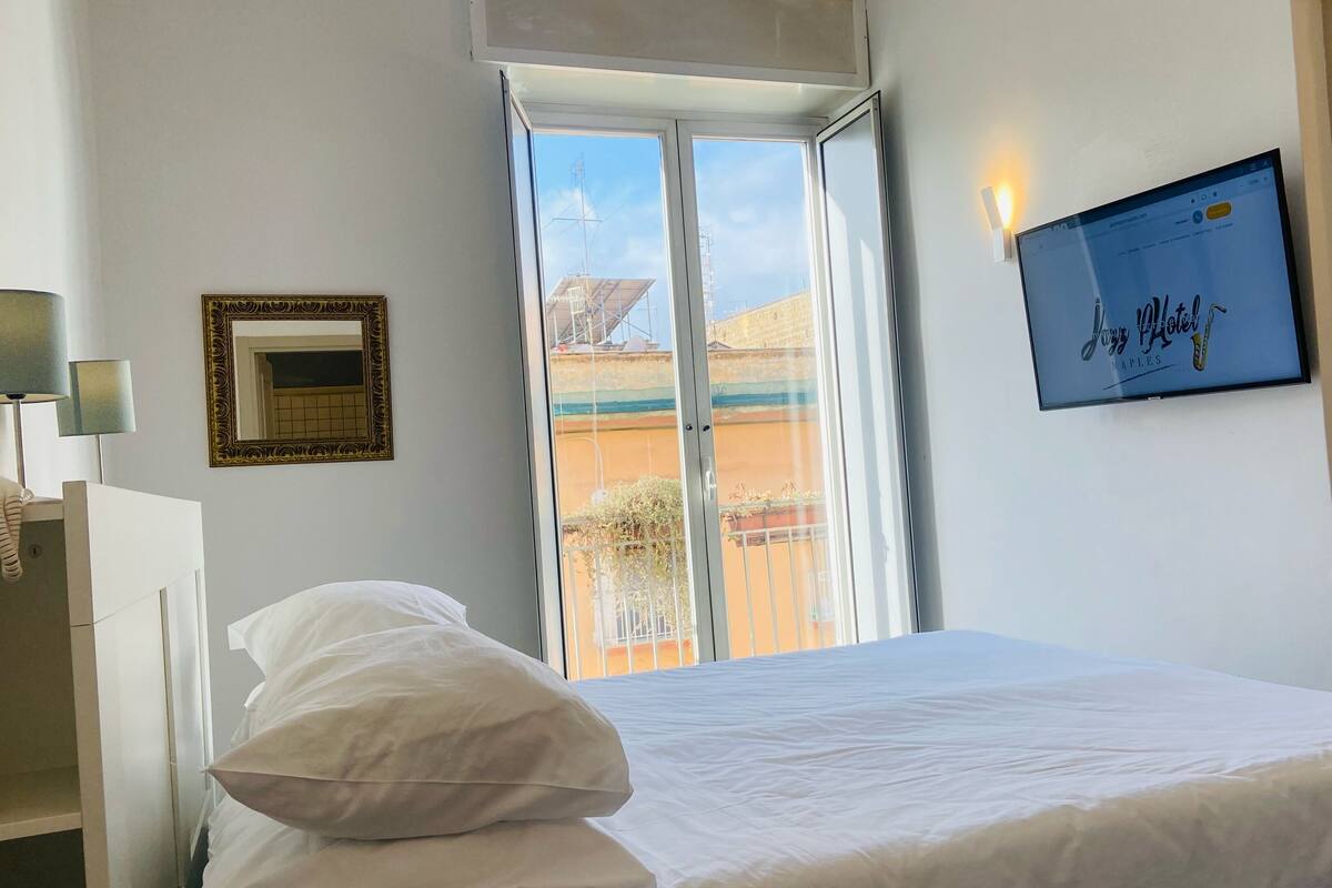 Une chambre d’hôtel avec un lit, une fenêtre donnant sur des immeubles, un tableau encadré et un téléviseur fixé au mur.