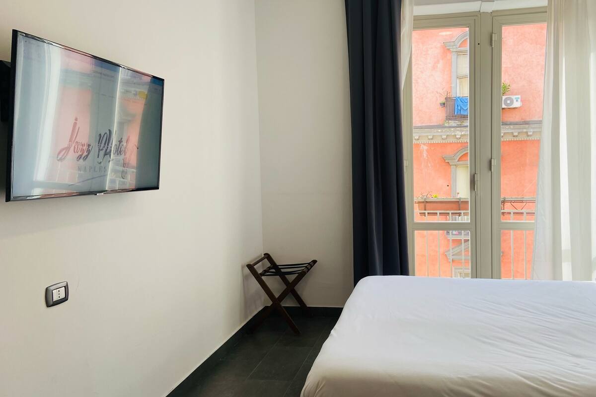 Une chambre d’hôtel avec un lit, une chaise, un téléviseur fixé au mur et une fenêtre avec des rideaux.