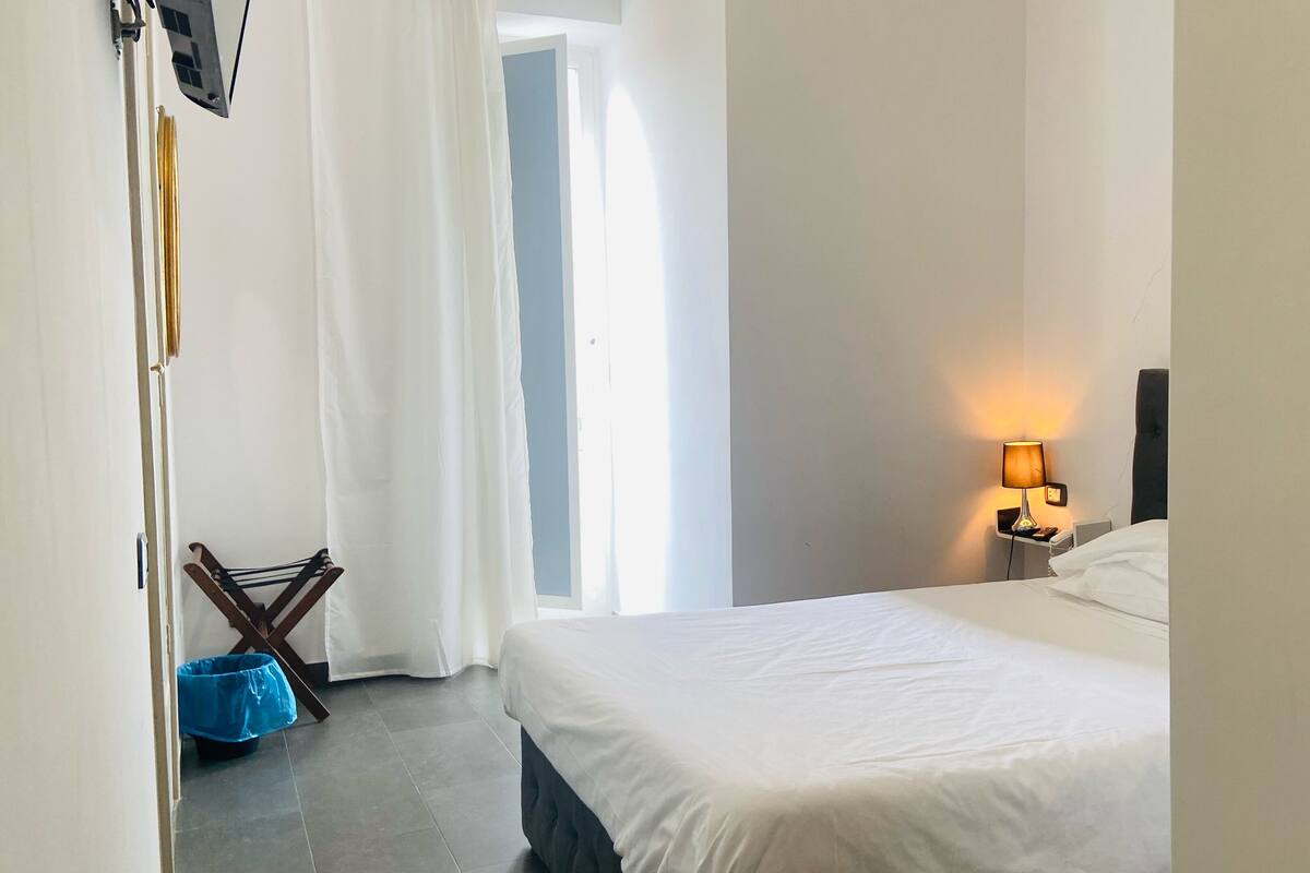Une chambre d’hôtel avec un lit, une table de chevet avec une lampe, un sac bleu et une porte donnant sur une autre pièce.