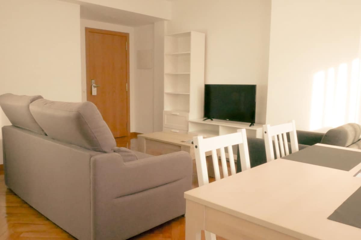 Un salon avec un canapé gris, une table blanche et un téléviseur posé sur un meuble.
