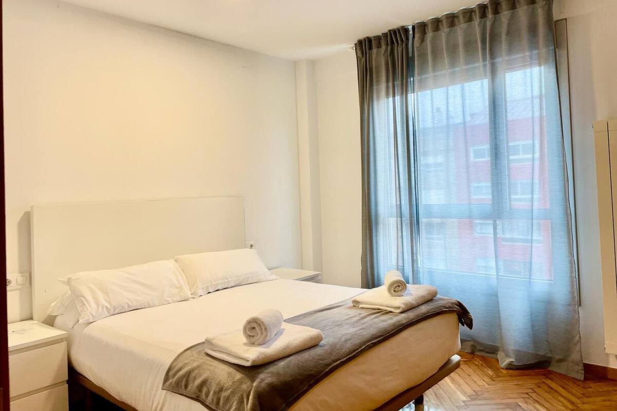 A hotel room with a large bed, a bedside table, a window with curtains, and a wooden floor.