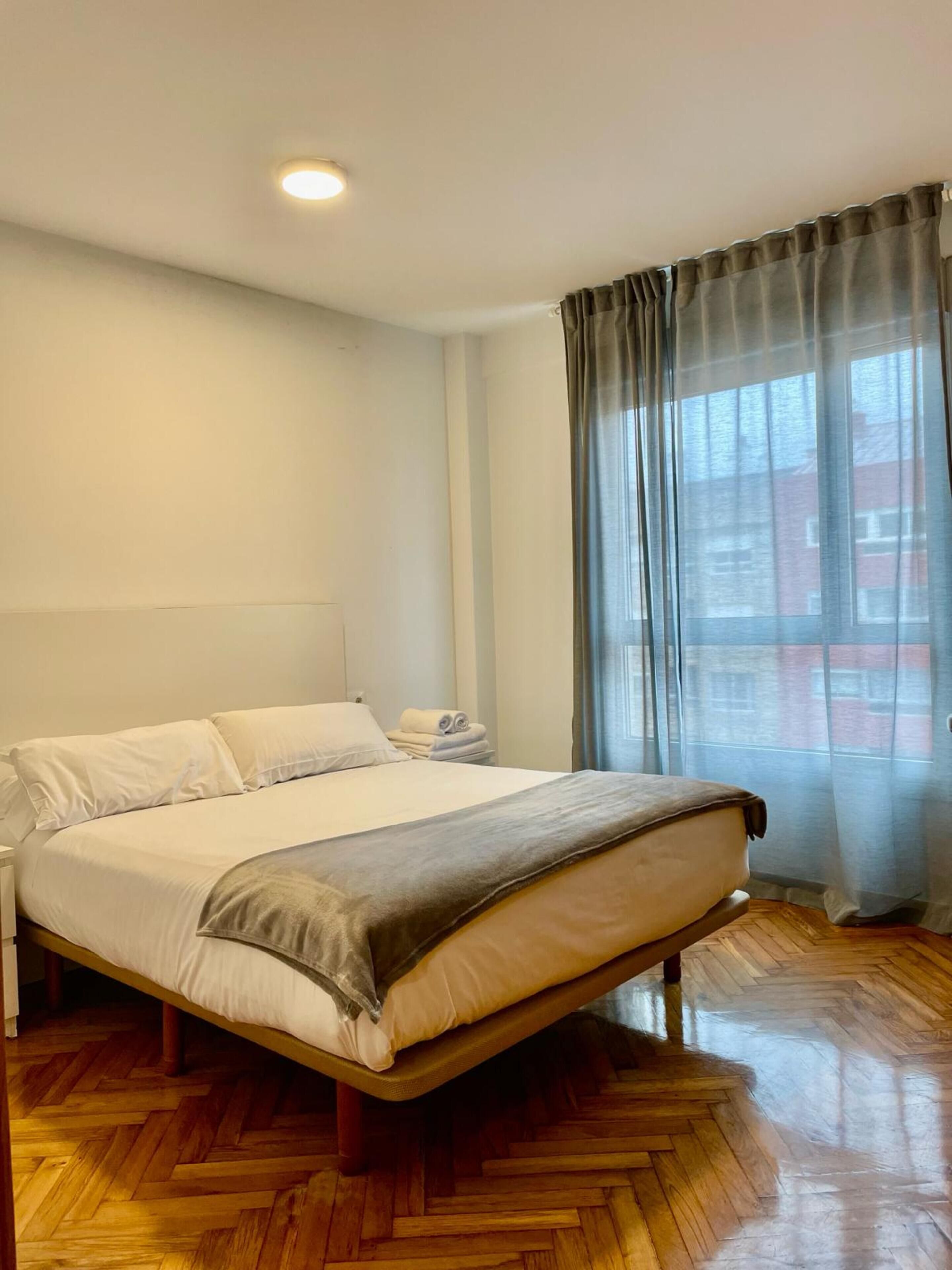A modern hotel room with a large bed, wooden flooring, a nightstand, and a window with curtains.