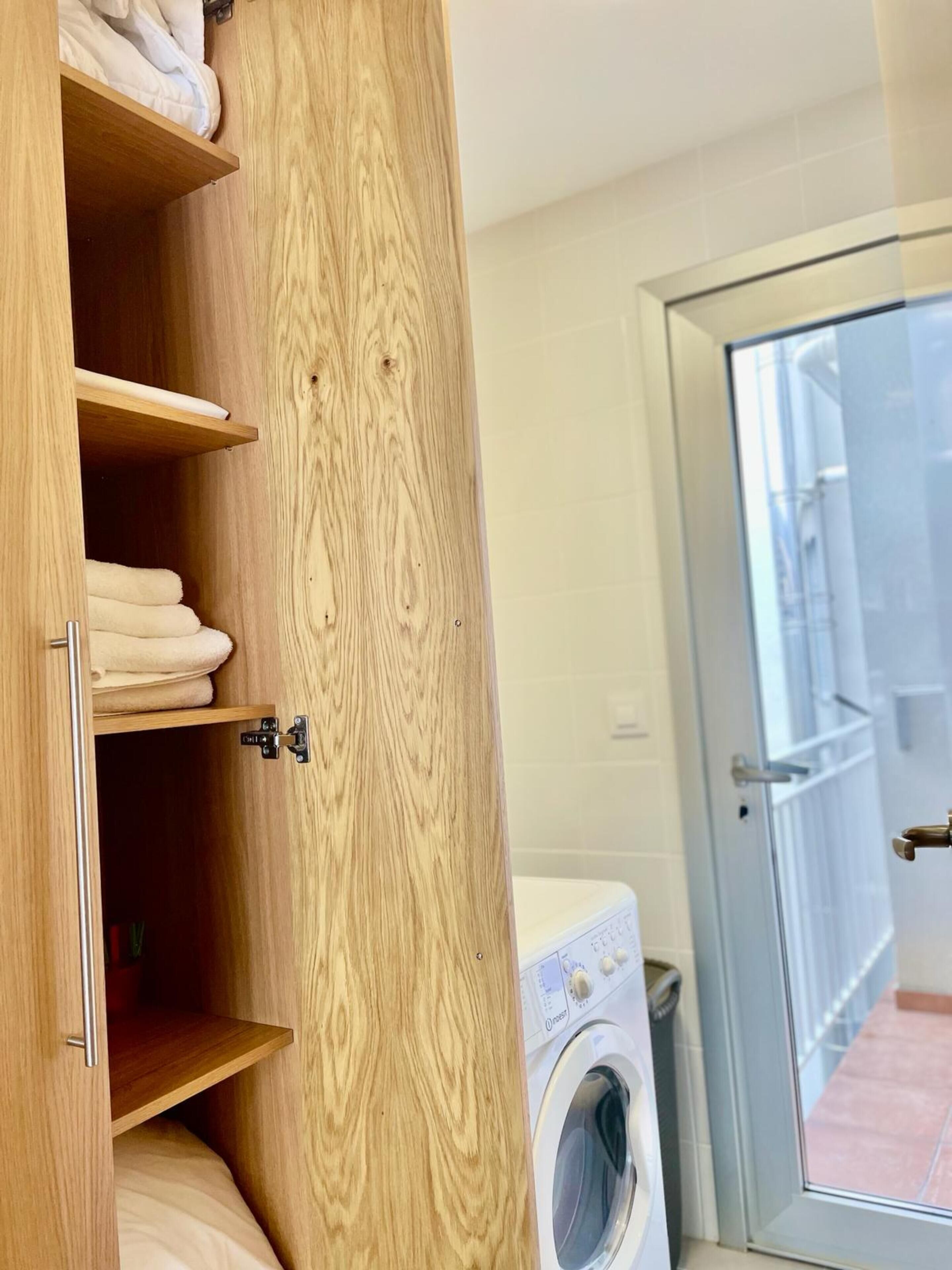 A wooden wardrobe with shelves and a hanging space, a washing machine, and a glass door leading to an outdoor area.