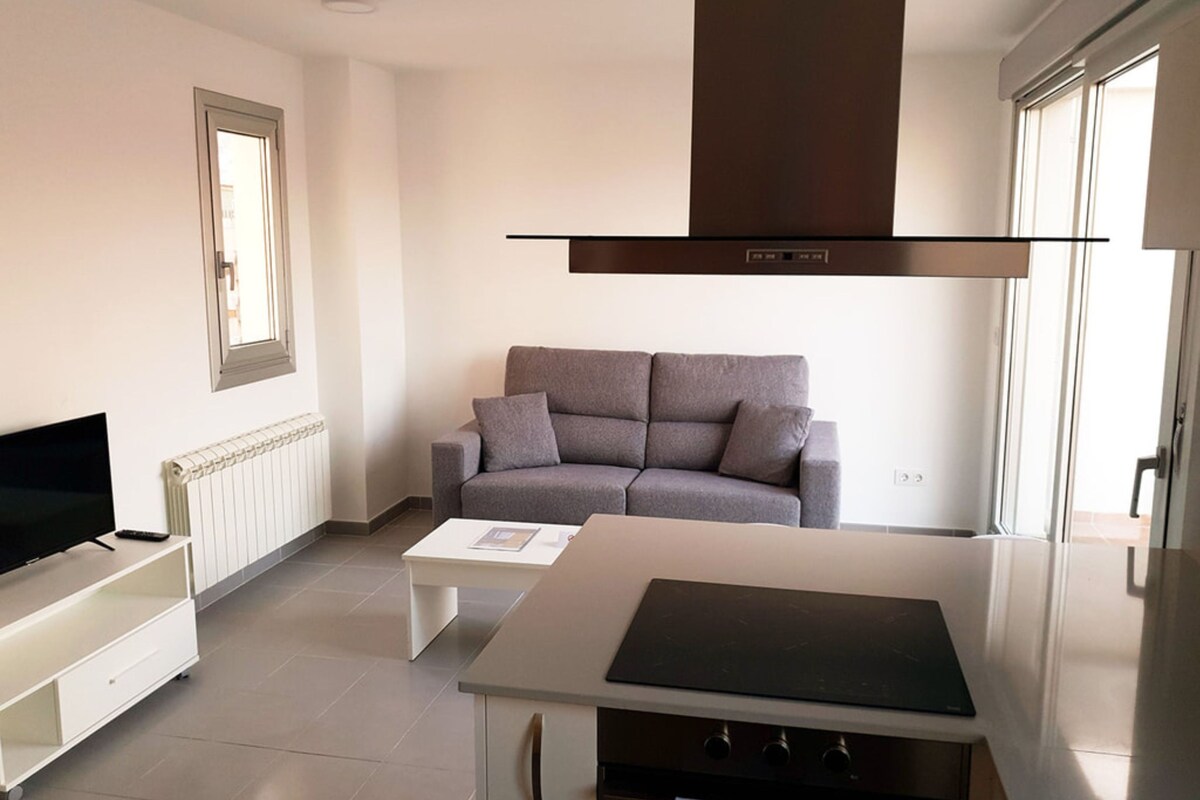 Una sala moderna con un sofá gris, un televisor de pantalla plana, una mesa de centro blanca y una cocina con estufa.