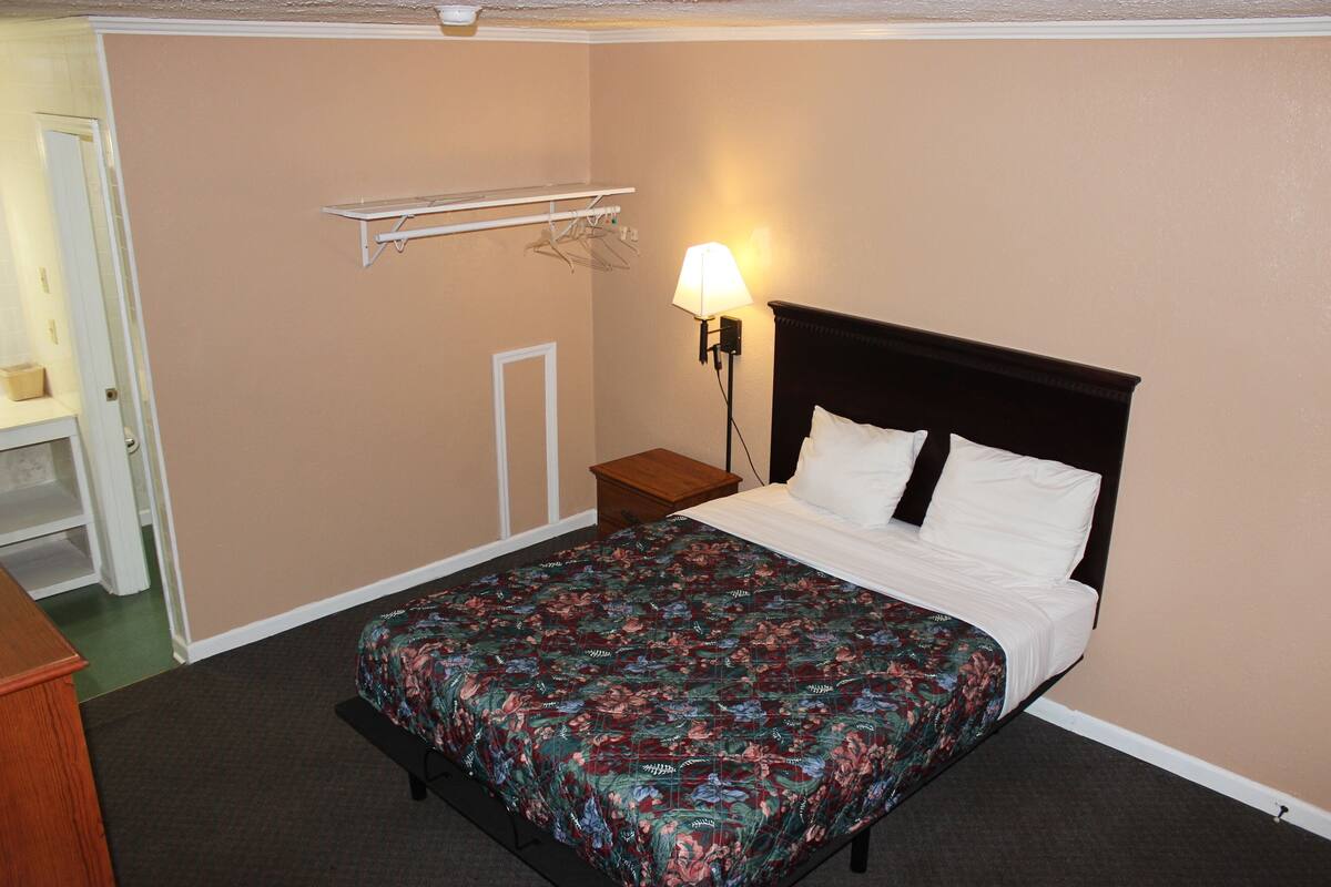 Quarto com cama, mesa de cabeceira, abajur, prateleira com roupas e uma porta.