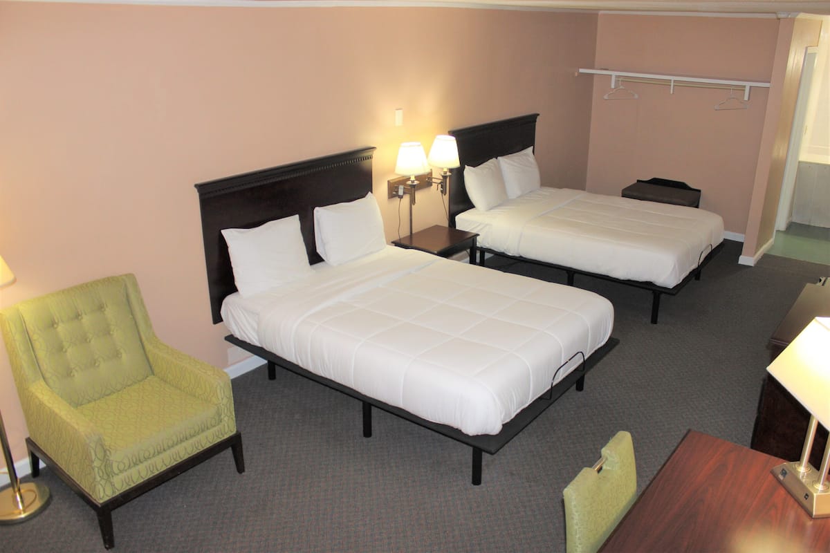 Quarto de hotel com duas camas, uma poltrona verde, mesa de cabeceira com luminárias e mesa de apoio de madeira.
