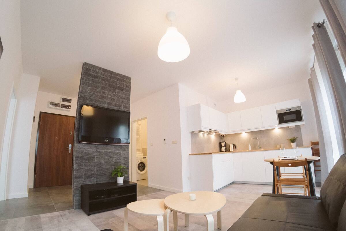 Ein modernes Wohnzimmer mit Flachbildfernseher, Kamin, Essbereich mit Tisch und Stühlen sowie Küche mit weißen Schränken und einer Mikrowelle.
