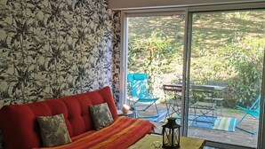 A room with a red sofa, patterned wallpaper, a wooden table, and a view of an outdoor deck with chairs and a table. - Biarritz charming two-room apartment with garden and parking! (Biarritz)