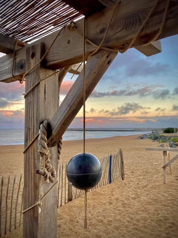 Una estructura de madera con cuerdas y un objeto esférico colgando, con vista a una playa al atardecer.