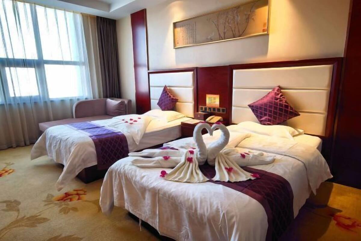 A hotel room with two beds, a sofa, a large window with curtains, and a wall-mounted artwork.