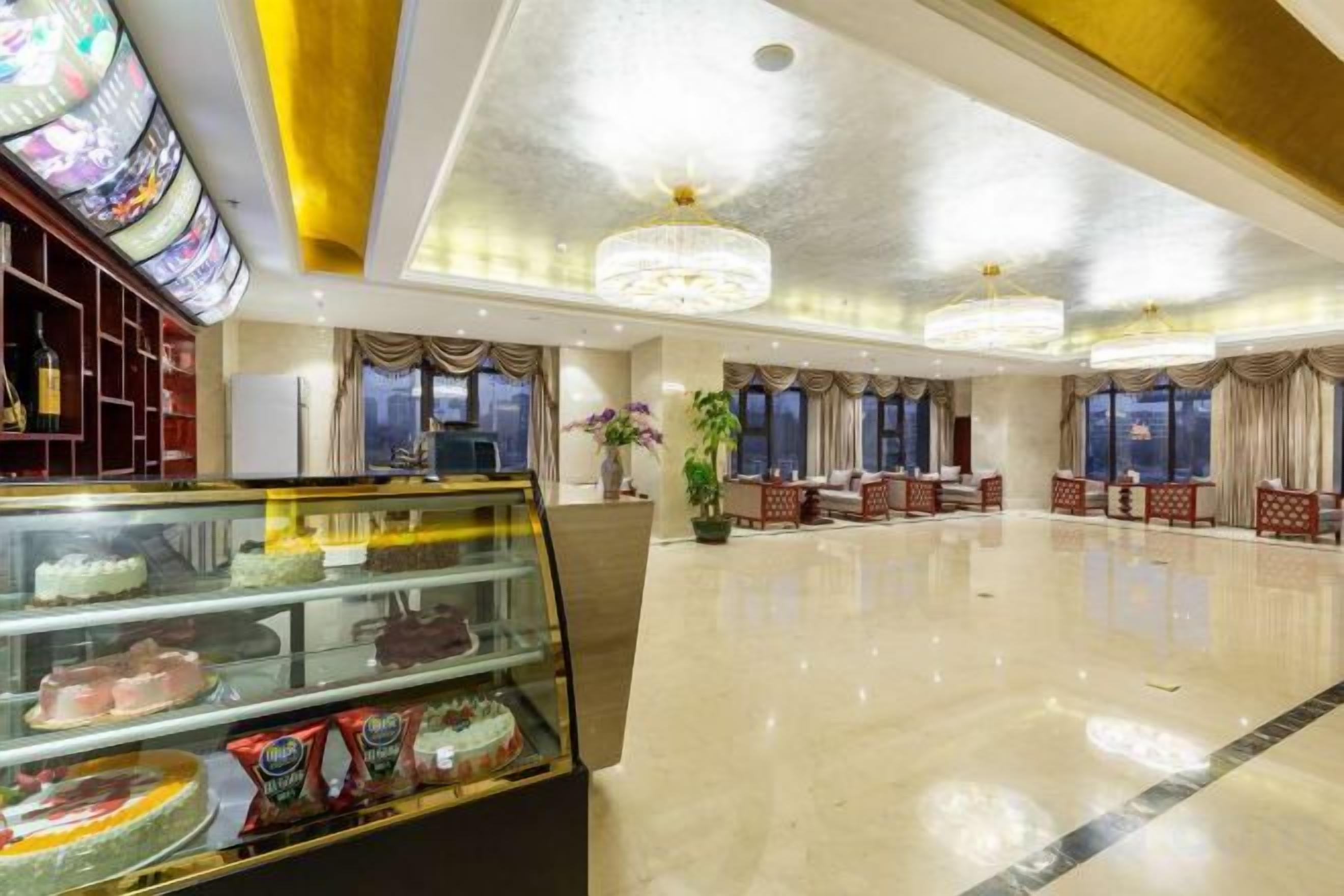 A hotel lobby with a glass display case of desserts, a chandelier, and a seating area with red upholstered chairs.
