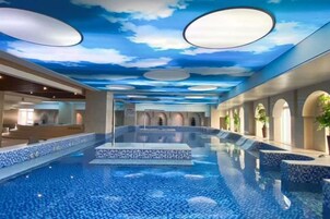Una amplia piscina interior con techo que imita un cielo nublado, rodeada de zonas de estar y un diseño arquitectónico moderno.