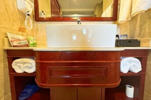 Un baño con lavamanos blanco, tocador de madera y repisas con toallas y artículos de aseo.