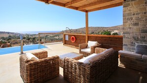 A covered outdoor seating area with wicker furniture, a stone wall, and a view of the landscape. - Phaestias Terra Villas (Faistos)
