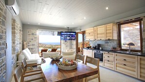 A modern kitchen with wooden cabinets, a dining area with a wooden table and chairs, and a stone wall. - Phaestias Terra Villas (Faistos)