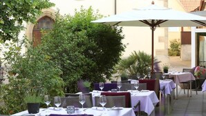 An outdoor dining area with tables and chairs, surrounded by greenery and a building with arched windows.