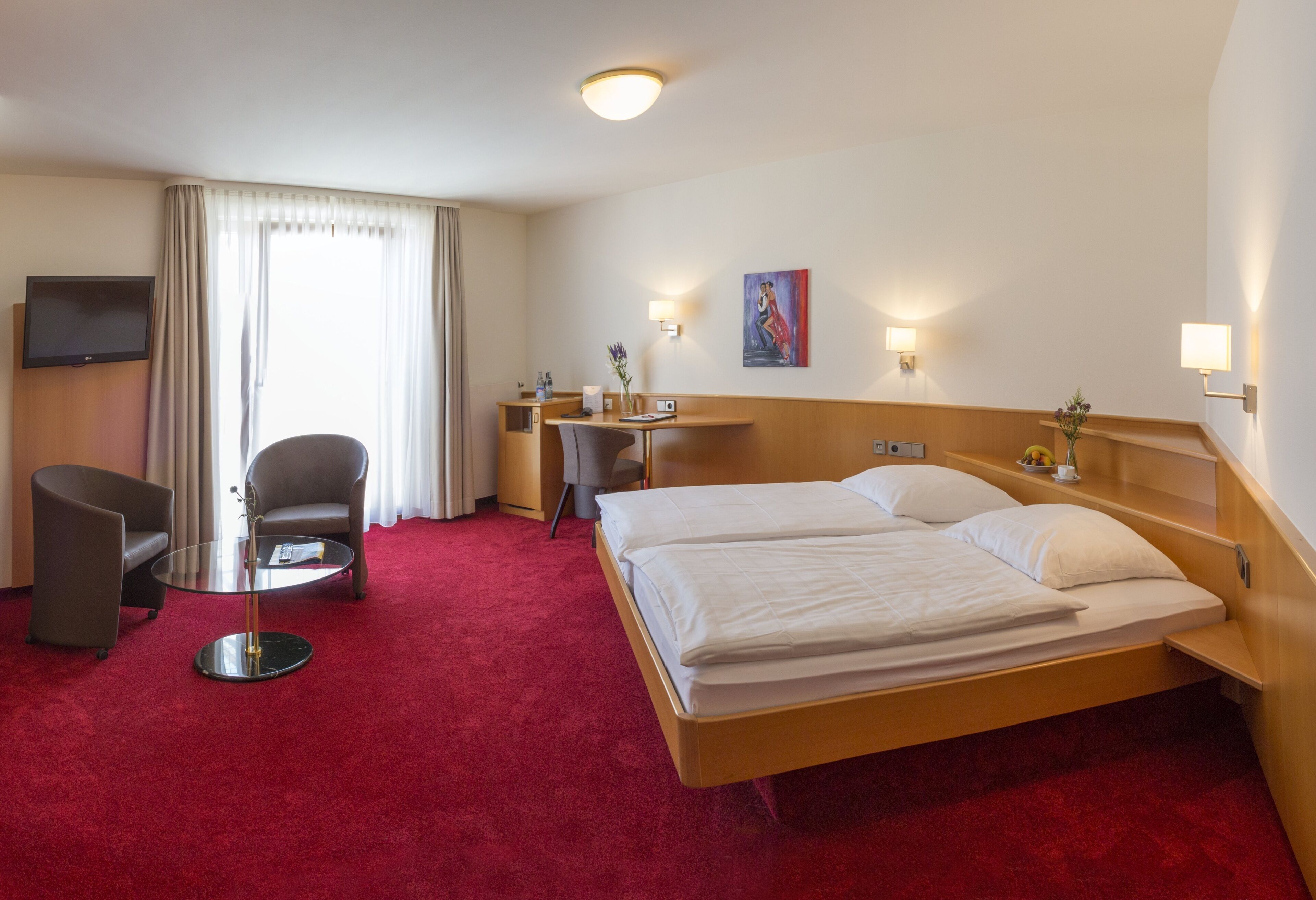 A hotel room with a large bed, a desk, a chair, a TV, and a painting on the wall.
