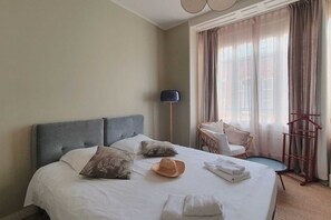 A hotel room with a large bed, a chair, a floor lamp, and a view of the outside through sheer curtains.