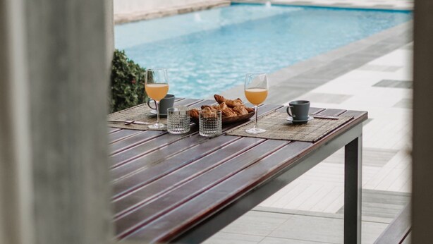 Une table de petit-déjeuner installée au bord de la piscine, avec des croissants, des verres de jus d’orange et une tasse de café.