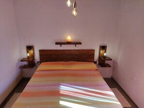A bedroom with a wooden headboard, a bed with a multicolored blanket, two bedside tables with lamps, and a wall-mounted shelf with a candle.