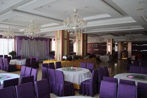 Een feestzaal met ronde tafels en paarse stoelen, kroonluchters en een podium.
