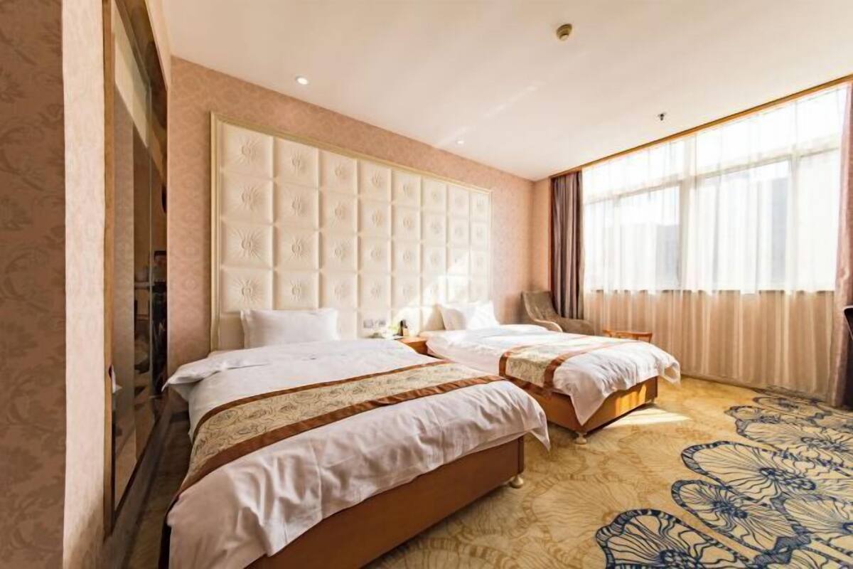 A hotel room with two beds, a large padded headboard, a patterned carpet, and a large window with curtains.