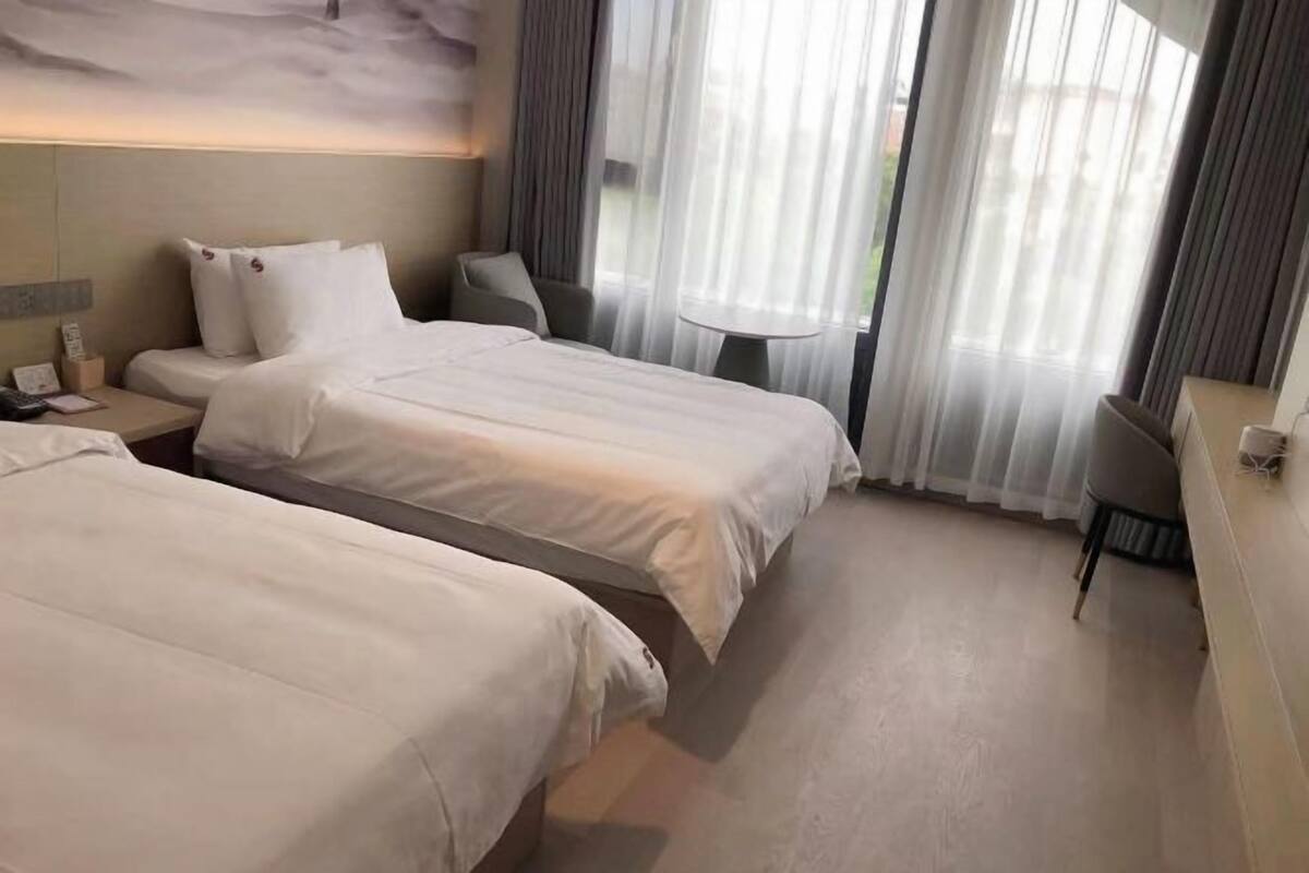 A hotel room with two beds, a desk, a chair, and a view of the outdoors through sheer curtains.