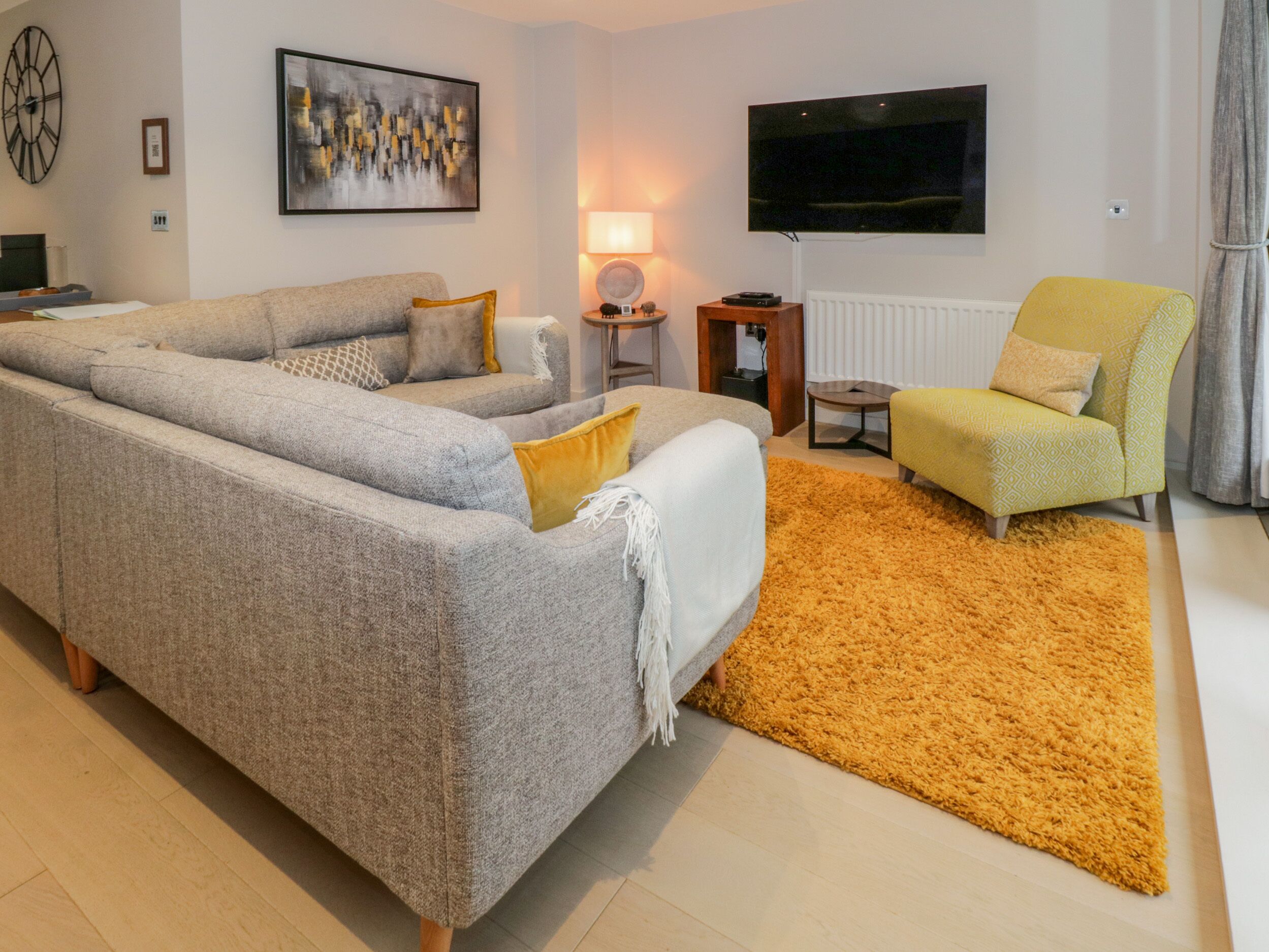 A modern living room with a grey sofa, a yellow armchair, a small round table, a lamp, and a flat-screen TV mounted on the wall.