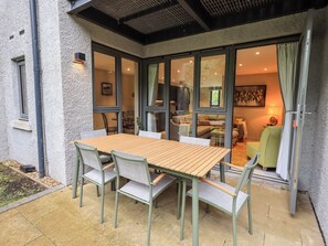 Eine Terrasse mit Holztisch und -stühlen, eine Glastür zu einem Innenraum mit Sofa und Lampe.