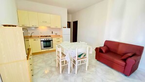 A compact living space with a kitchenette, a dining table with chairs, and a red sofa. - Casa Vacanza Silvaredda (San Teodoro)