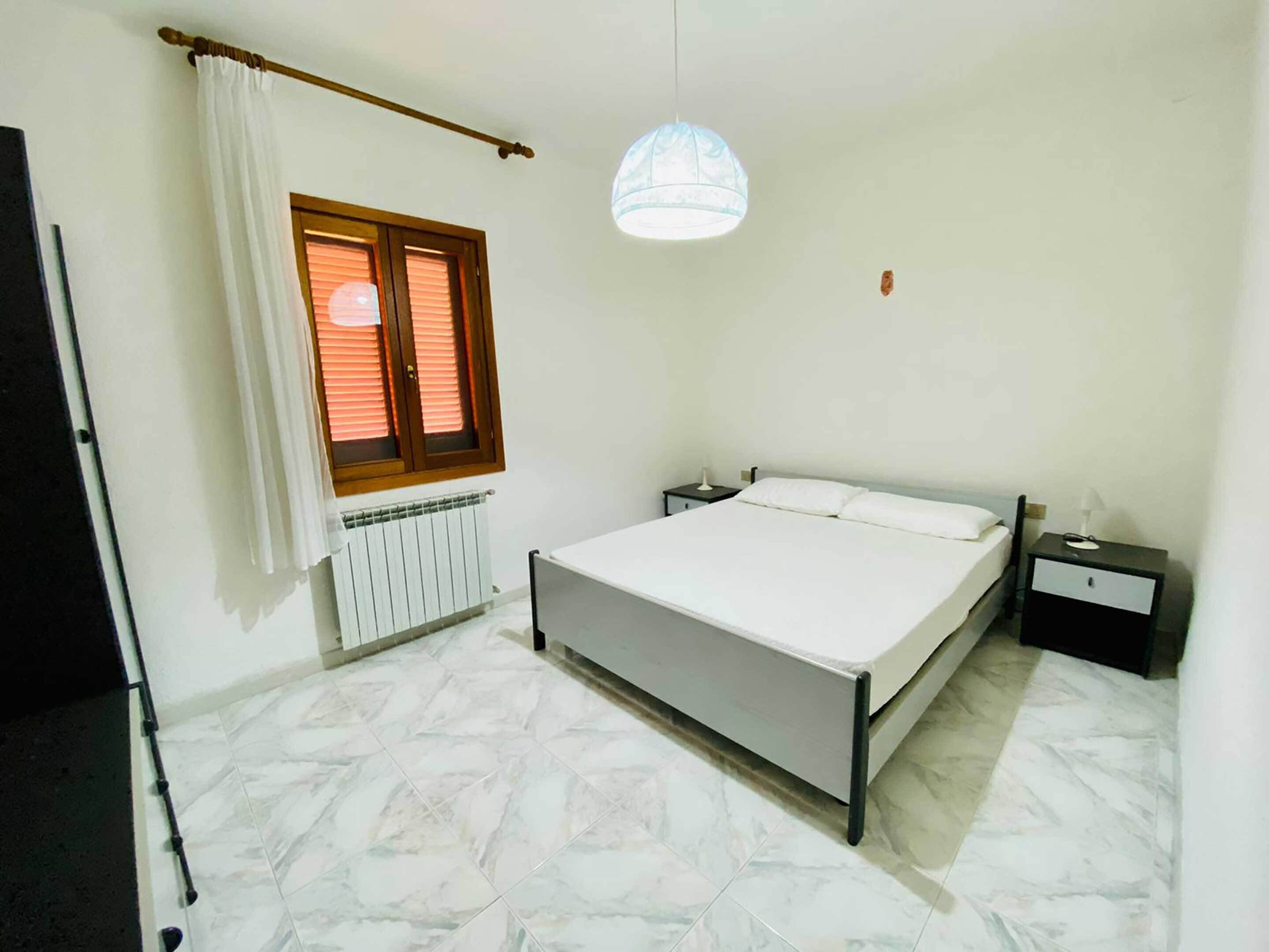 Une chambre à coucher comprenant un lit, une table de chevet, une lampe, une fenêtre avec des stores, un radiateur et un luminaire au plafond.
