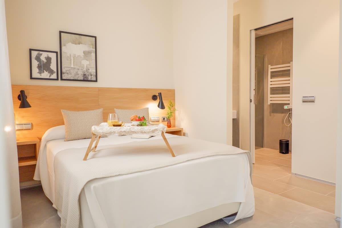 Una cama bien hecha con una bandeja de desayuno, una mesita de noche con lámpara y dos cuadros en la pared.