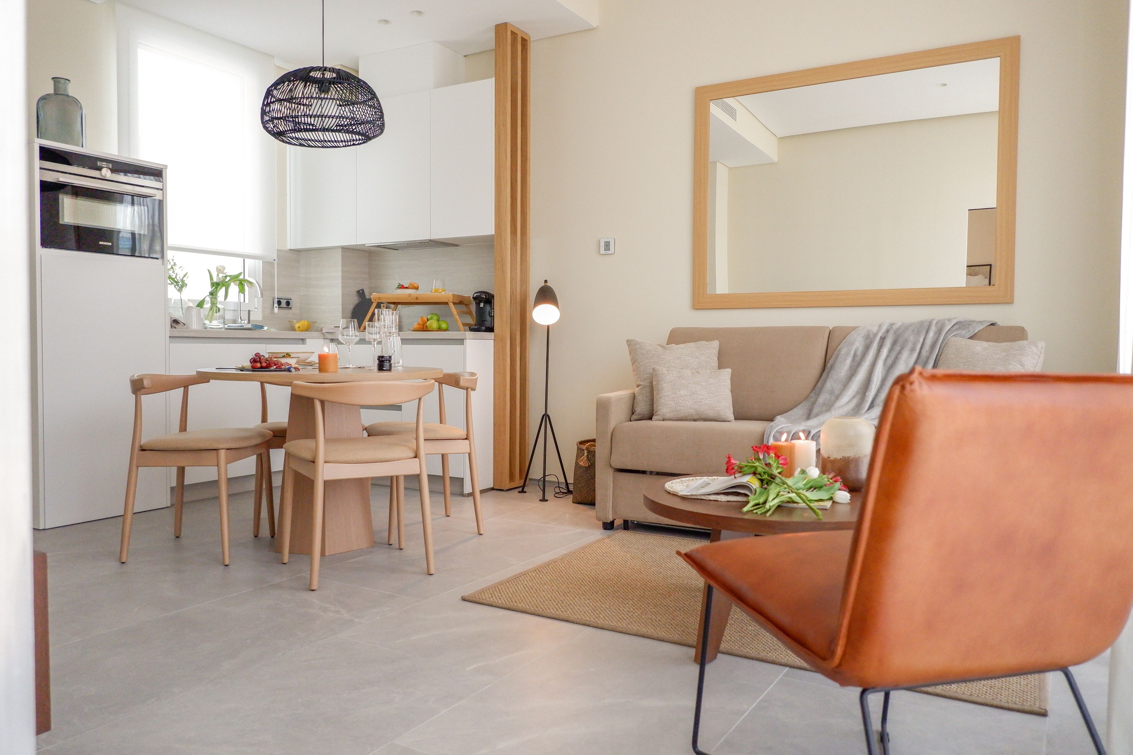 A modern kitchen with white cabinets, a dining area with wooden chairs, a sofa, and a large mirror on the wall.