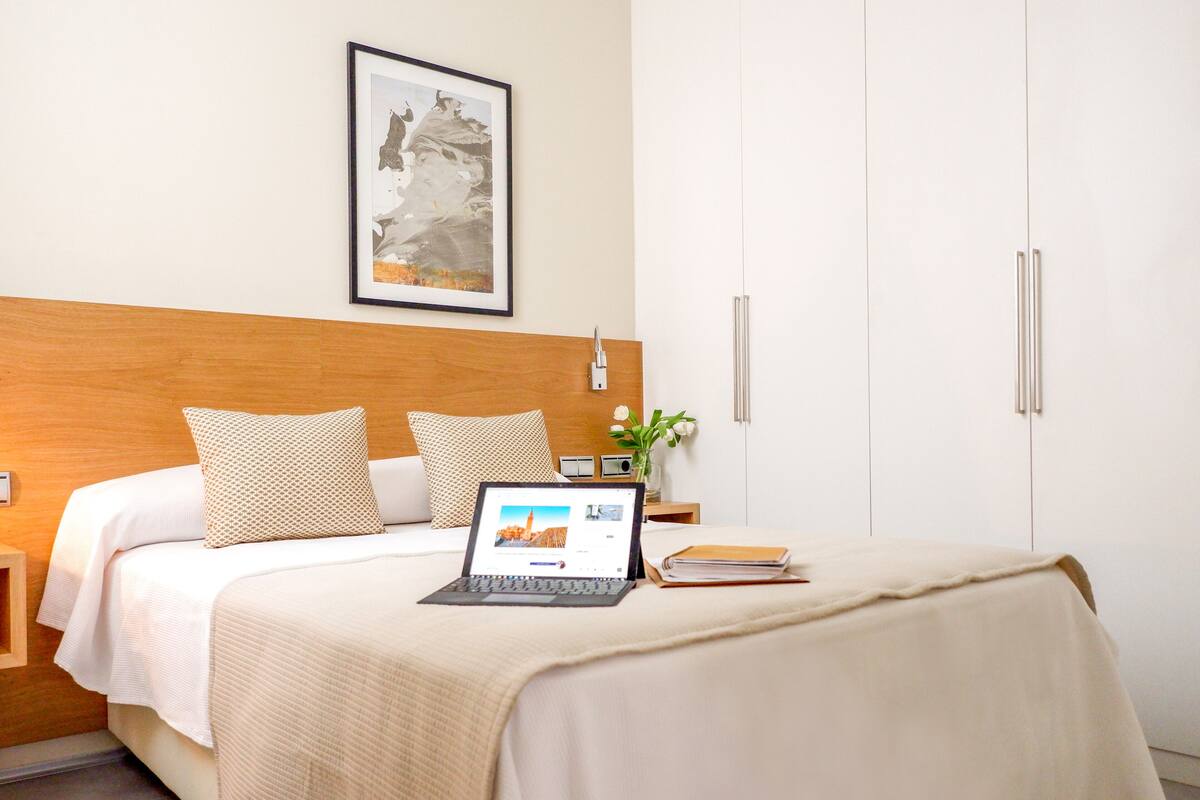 Una cama bien hecha con una computadora portátil, un cuadro en la pared y una mesita de noche con un florero.
