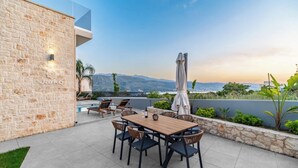 An outdoor patio with a stone wall, a wooden table, and chairs. There is a pool in the background with a view of mountains. - Villa Jarko (Chania)