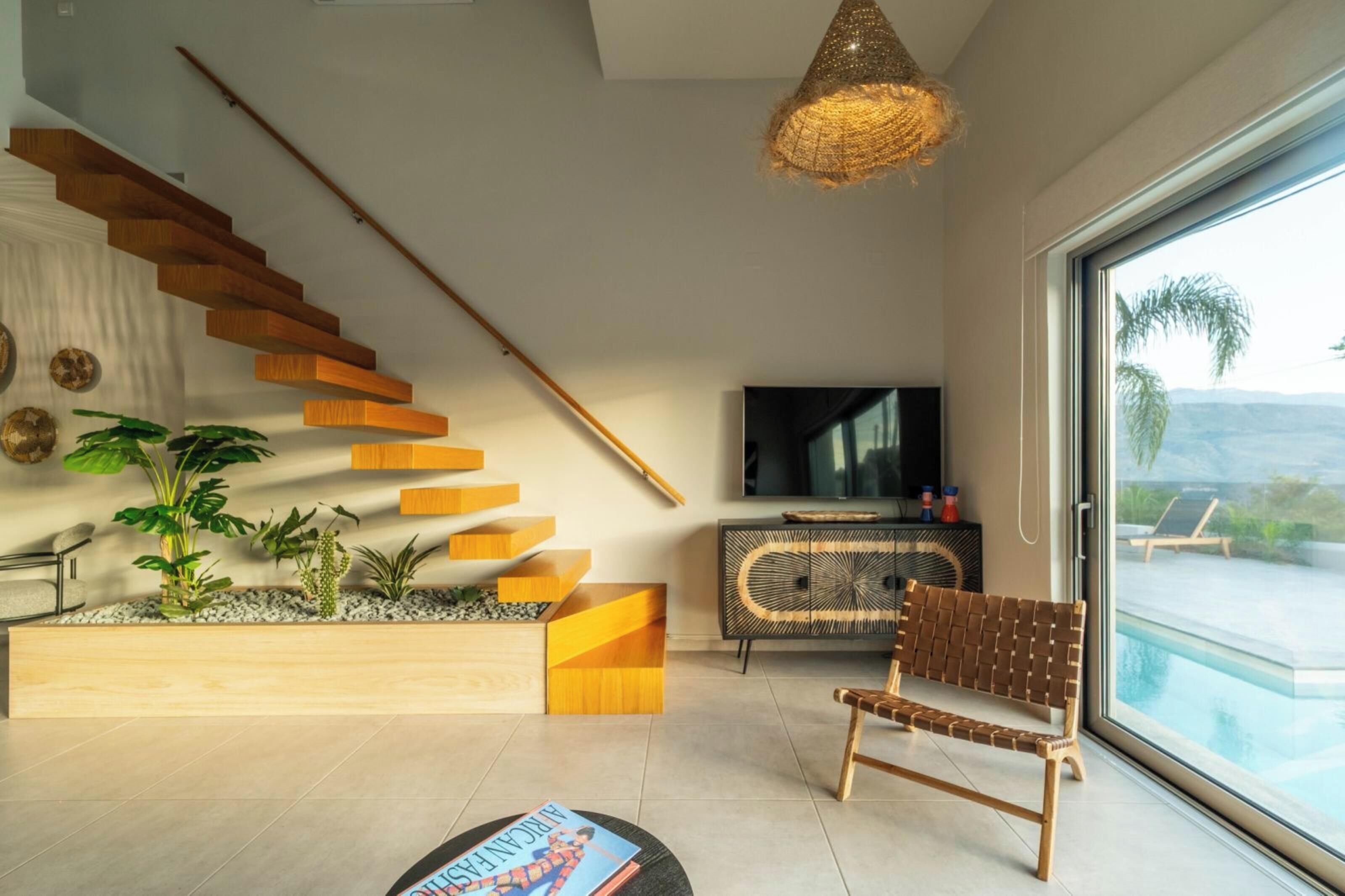 Un salon moderne avec un escalier en bois, un téléviseur à écran plat, un fauteuil en osier et une vue sur une piscine.