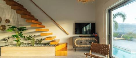 Un salon moderne avec un escalier en bois, un téléviseur à écran plat, un fauteuil en osier et une vue sur une piscine.
