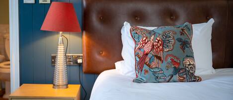 Una cama bien hecha con una almohada estampada, una lámpara roja sobre una mesita de noche de madera y una pared azul con un enchufe.