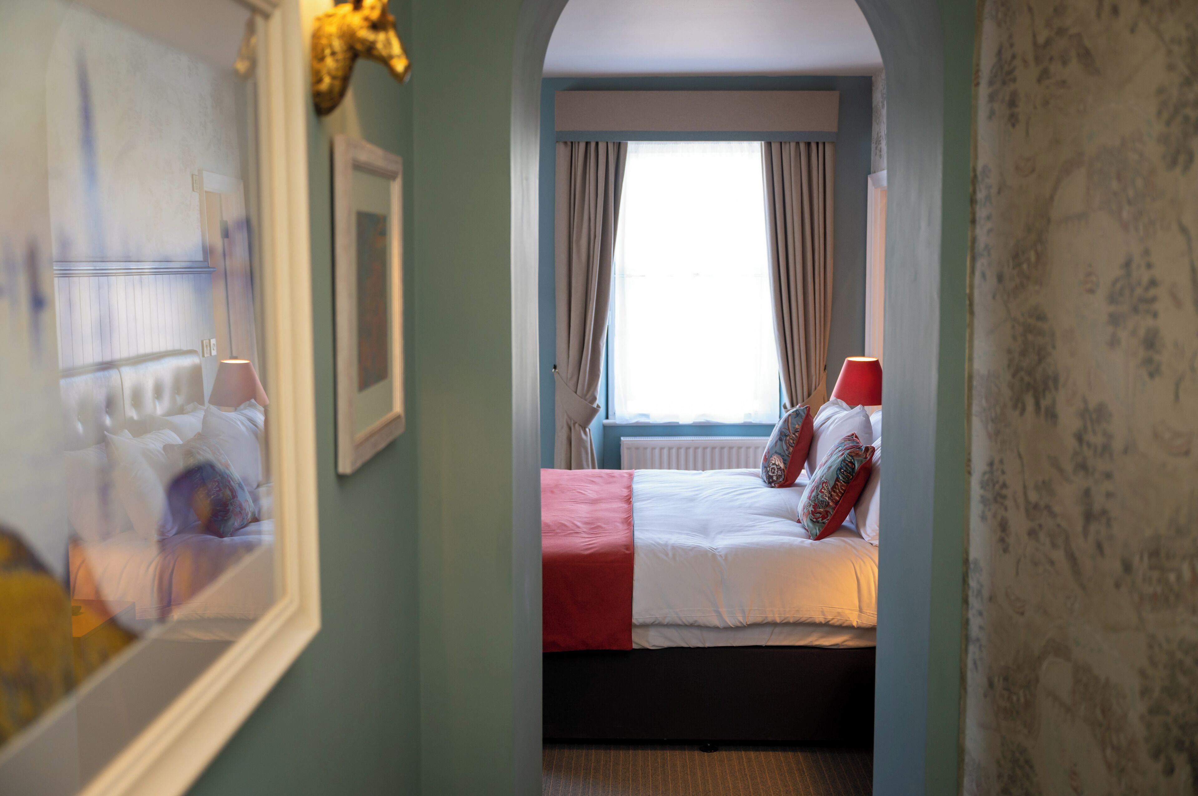 A bedroom with a bed, a window with curtains, and a hallway with a mirror.