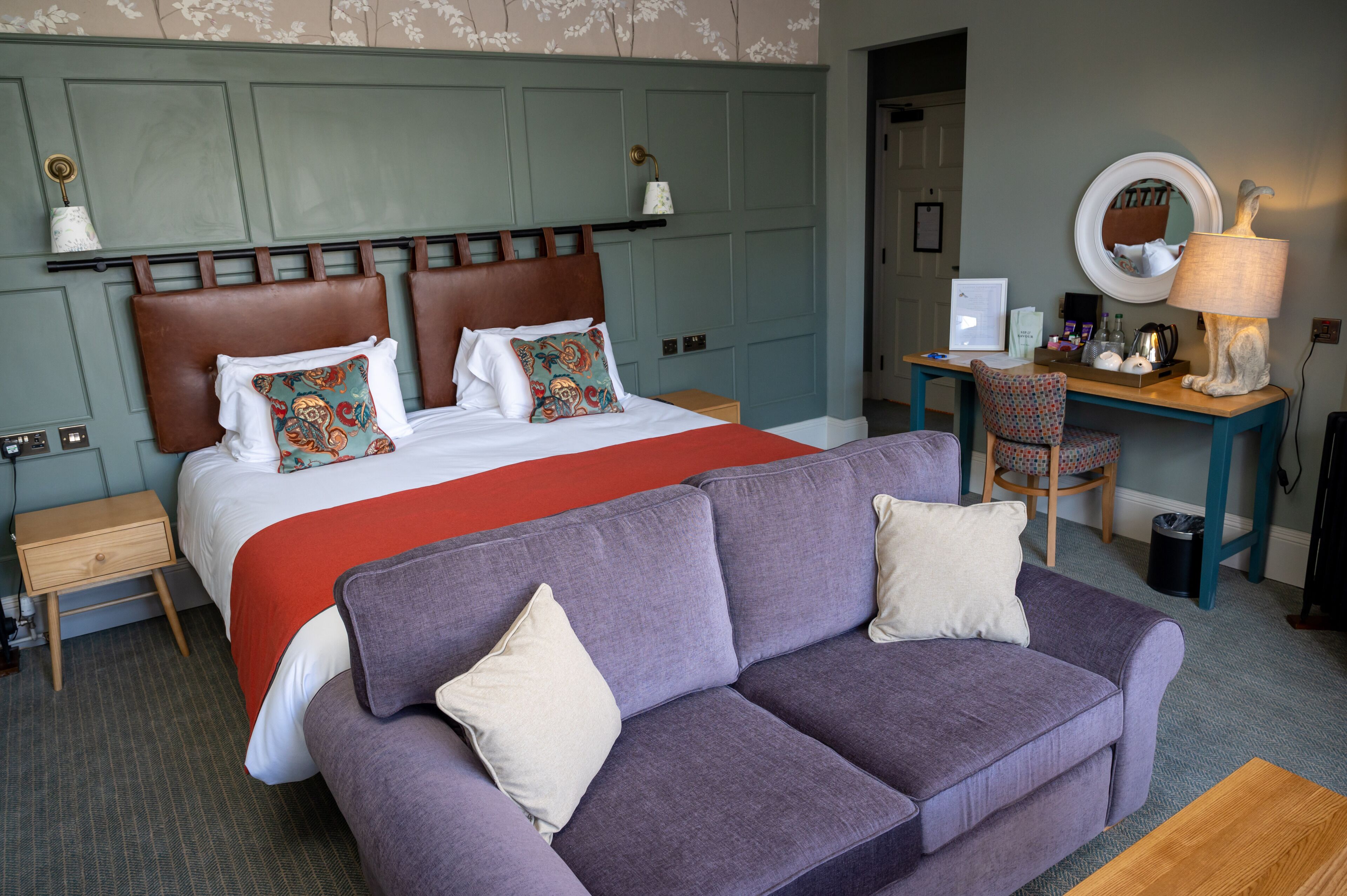 A hotel room with a large bed, a purple sofa, a desk with a computer, and a lamp.