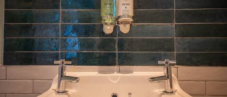 Un baño con lavamanos blanco, dos dispensadores de jabón para manos y una salpicadera de azulejos.
