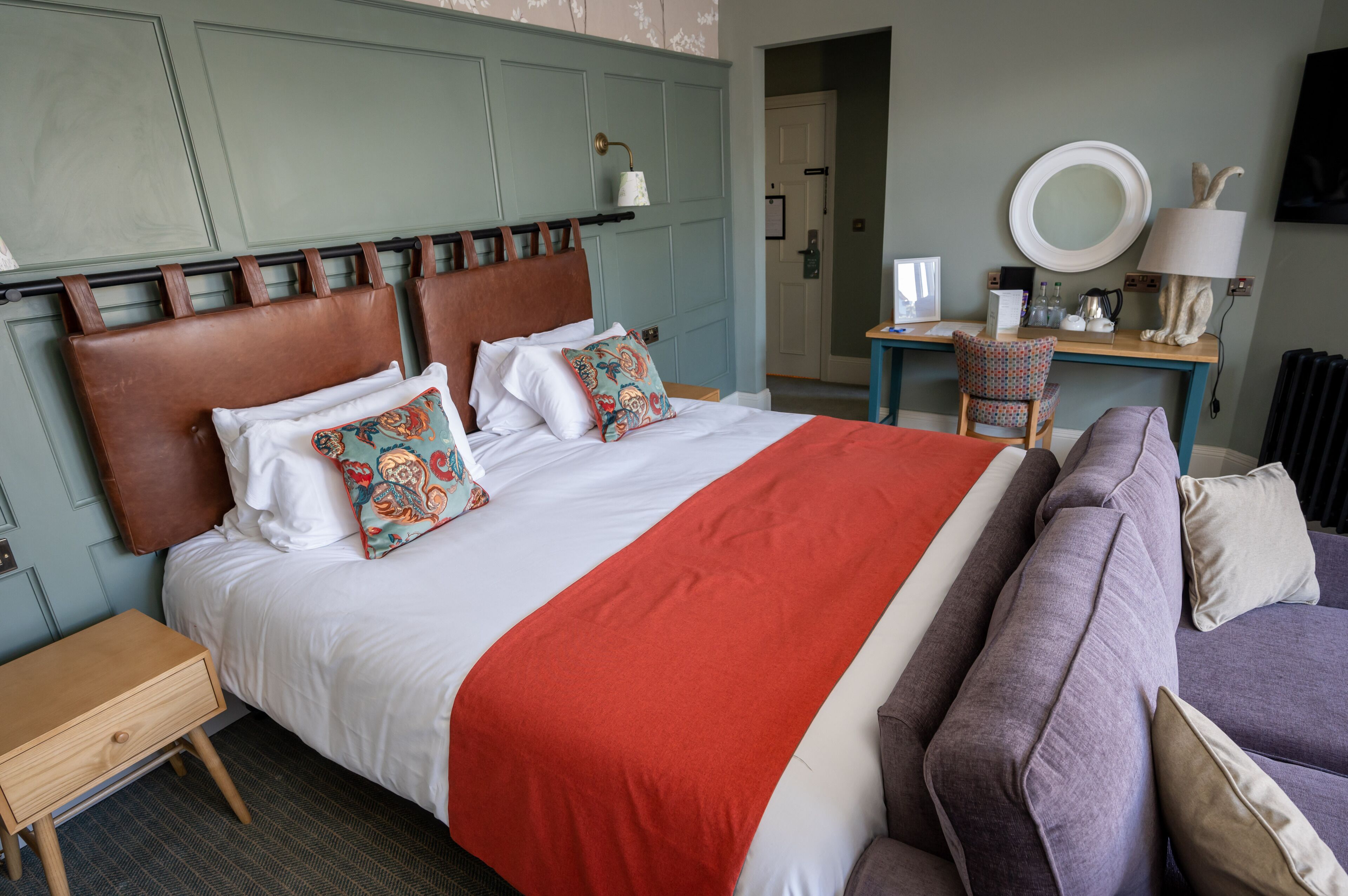 Een net opgemaakt bed met een rode deken, een bruin hoofdbord en decoratieve kussens. Er is een klein houten nachtkastje naast het bed, een paarse bank met kussens en een bureau met een lamp en een spiegel.