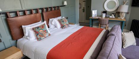 A neatly made bed with a red blanket, a brown headboard, and decorative pillows. There is a small wooden nightstand beside the bed, a purple sofa with cushions, and a desk with a lamp and a mirror.
