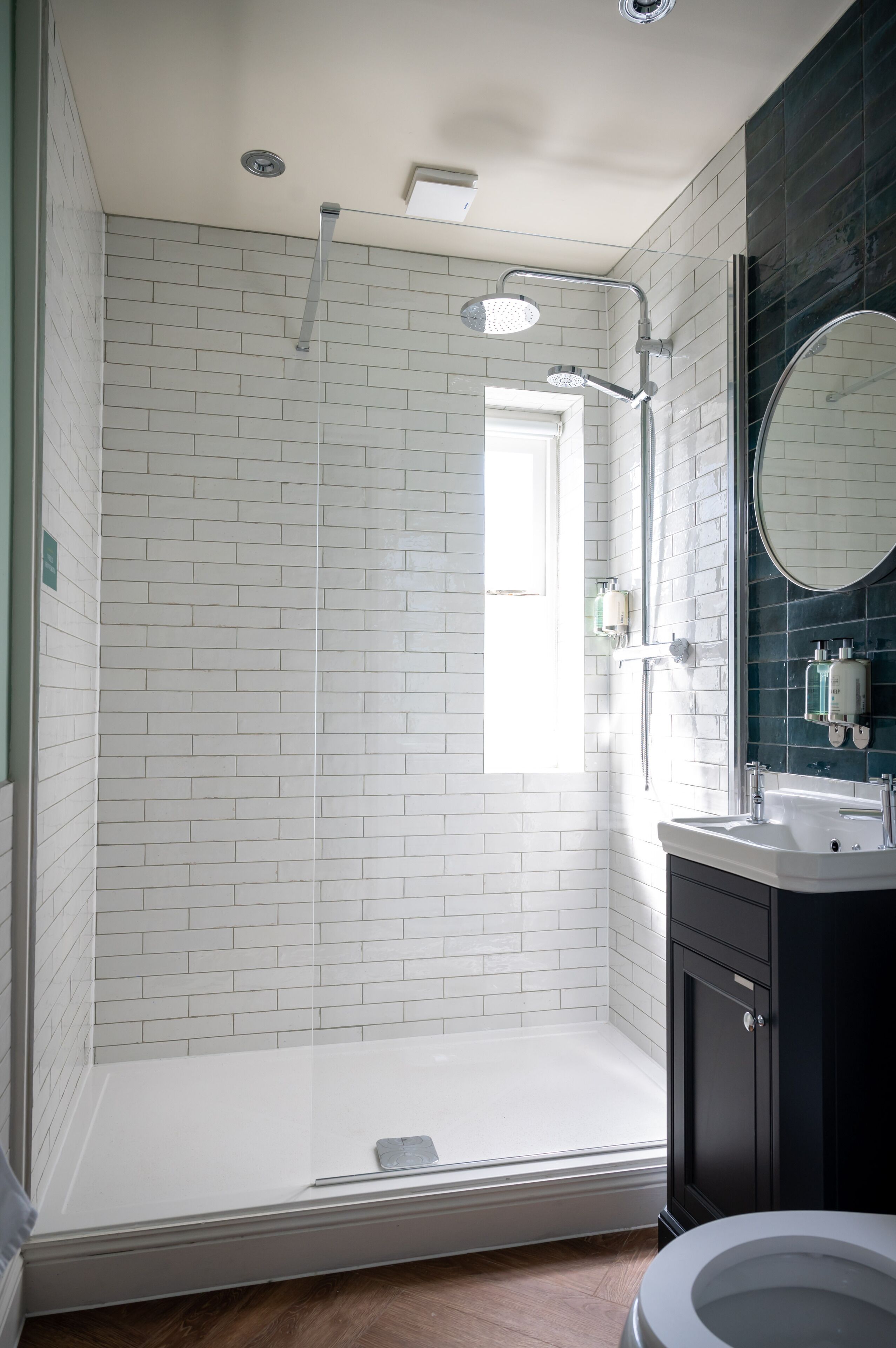A modern bathroom with white subway tiles, a glass shower enclosure, a black vanity with a round mirror, and a toilet.